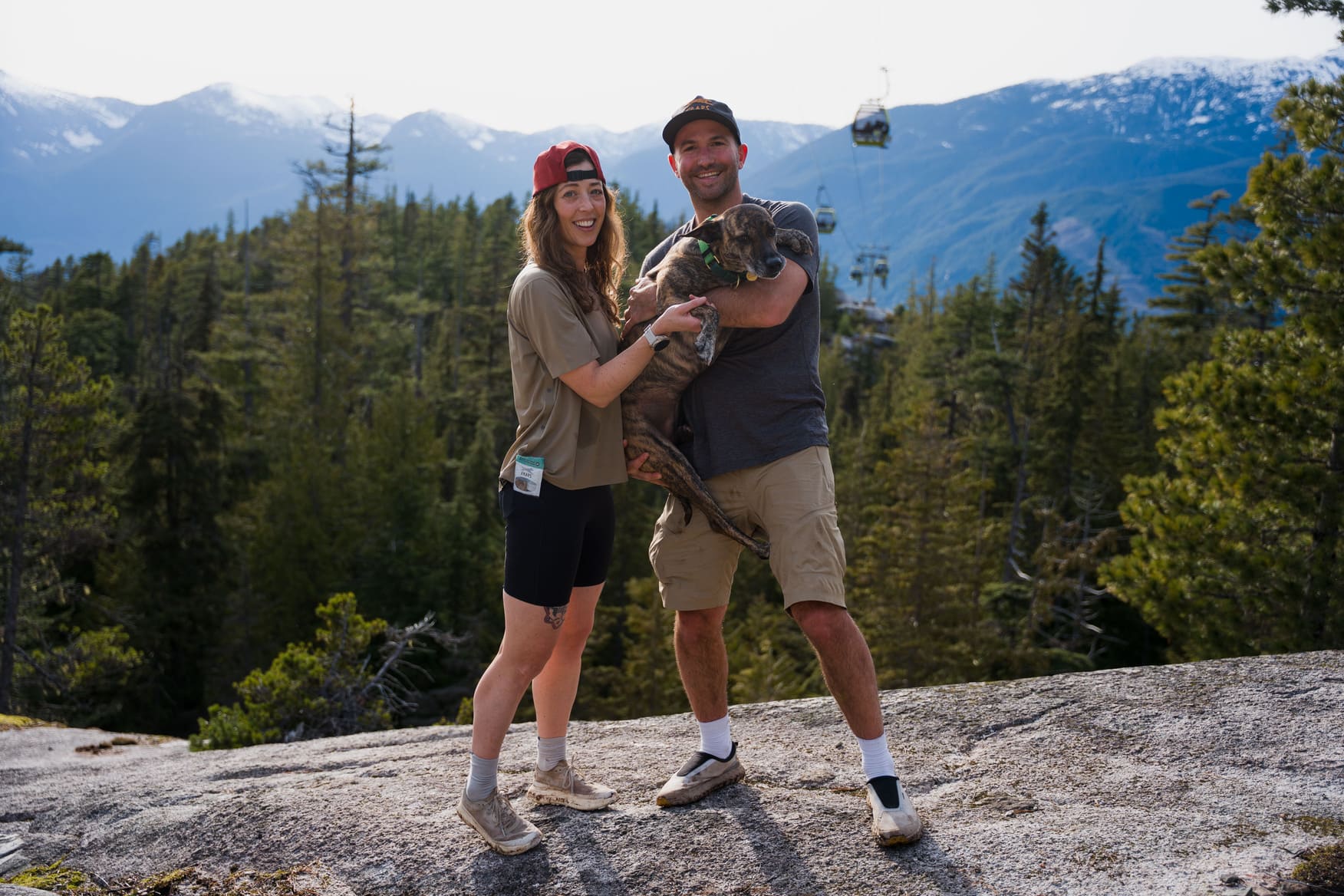 Two people with a dog on a mountain top with trees and mountains in the background